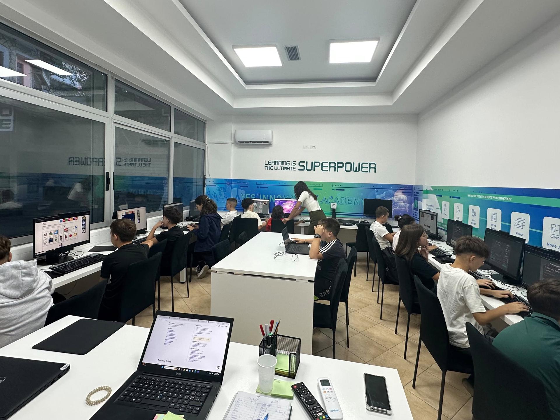 Students at their computers in the classroom, with a teacher helping at the back
